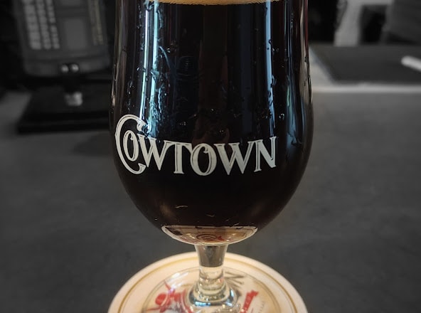 A glass of dark beer is displayed on a coaster, showcasing the engraved logo 'COWTOWN'. The beverage bubbles lightly, indicating freshness, and the smooth surface of the table reflects the glass. The blurred background suggests a social or lively environment.