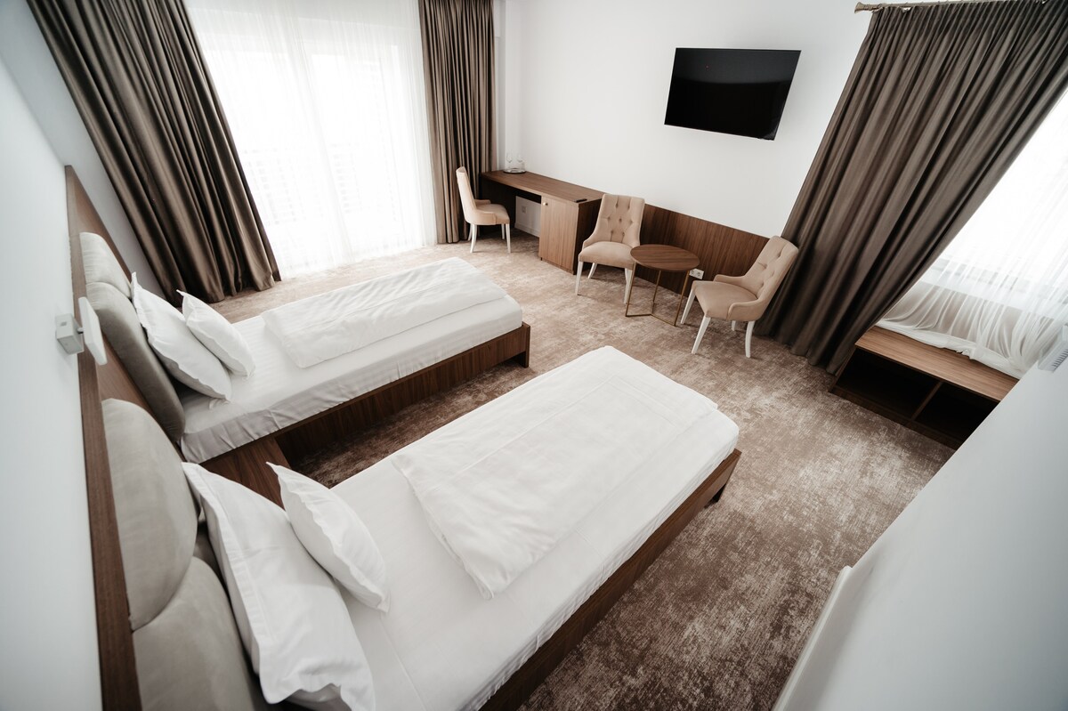 The sleeping area features two spacious beds with light-colored linens. A flat-screen television is mounted on the wall. A small desk and two chairs are positioned nearby. Natural light filters through sheer curtains, enhancing the airy feel of the room.