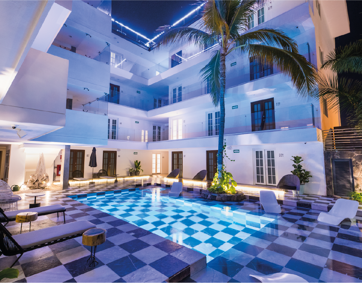 A serene courtyard is surrounded by a three-story building featuring multiple balconies and large windows. The center is dominated by a swimming pool with a checkered pattern, illuminated by soft lighting. Tropical plants and lounge furniture are positioned around the pool, enhancing relaxation.