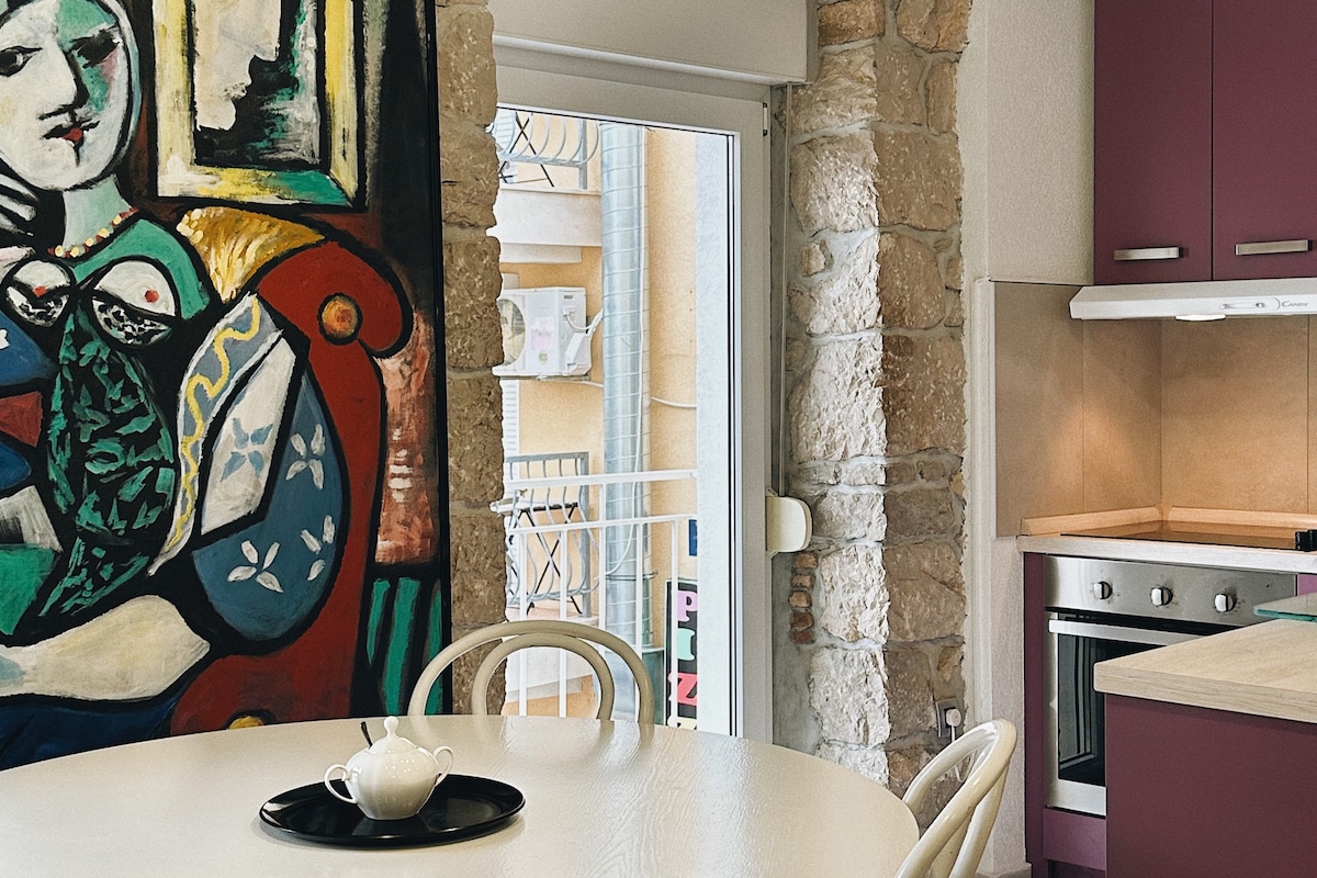 A modern kitchen area is highlighted by a round table with a black tray featuring a tea set. The space is adorned with a colorful artwork against the stone wall, and a window allows natural light to enter, showcasing a glimpse of the outside balcony.