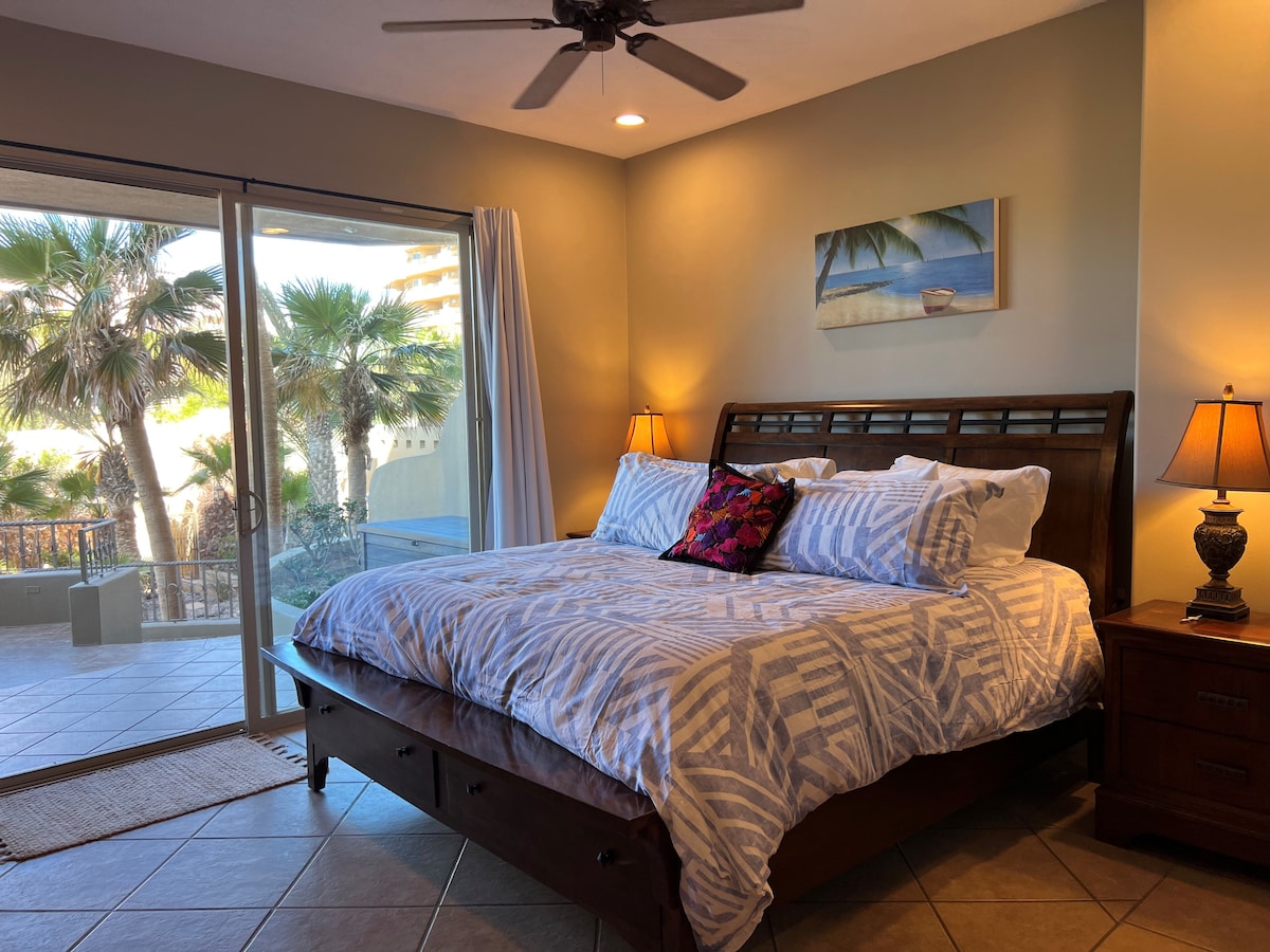 A spacious master bedroom is equipped with a king bed featuring luxurious bedding and decorative pillows. Natural light fills the room through large sliding glass doors, which provide views of lush palm trees and outdoor space. Two bedside lamps add soft illumination.