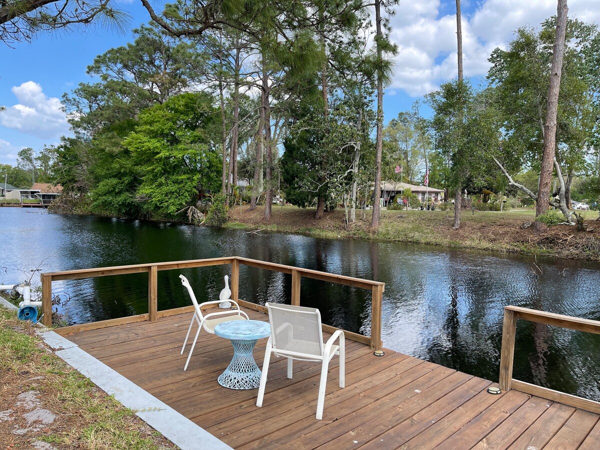A wooden deck extends over calm water, furnished with two white chairs and a small blue table. Lush greenery lines the opposite bank, while trees provide shade and a serene backdrop. The reflection of the trees is visible on the water's surface, enhancing the tranquil setting.