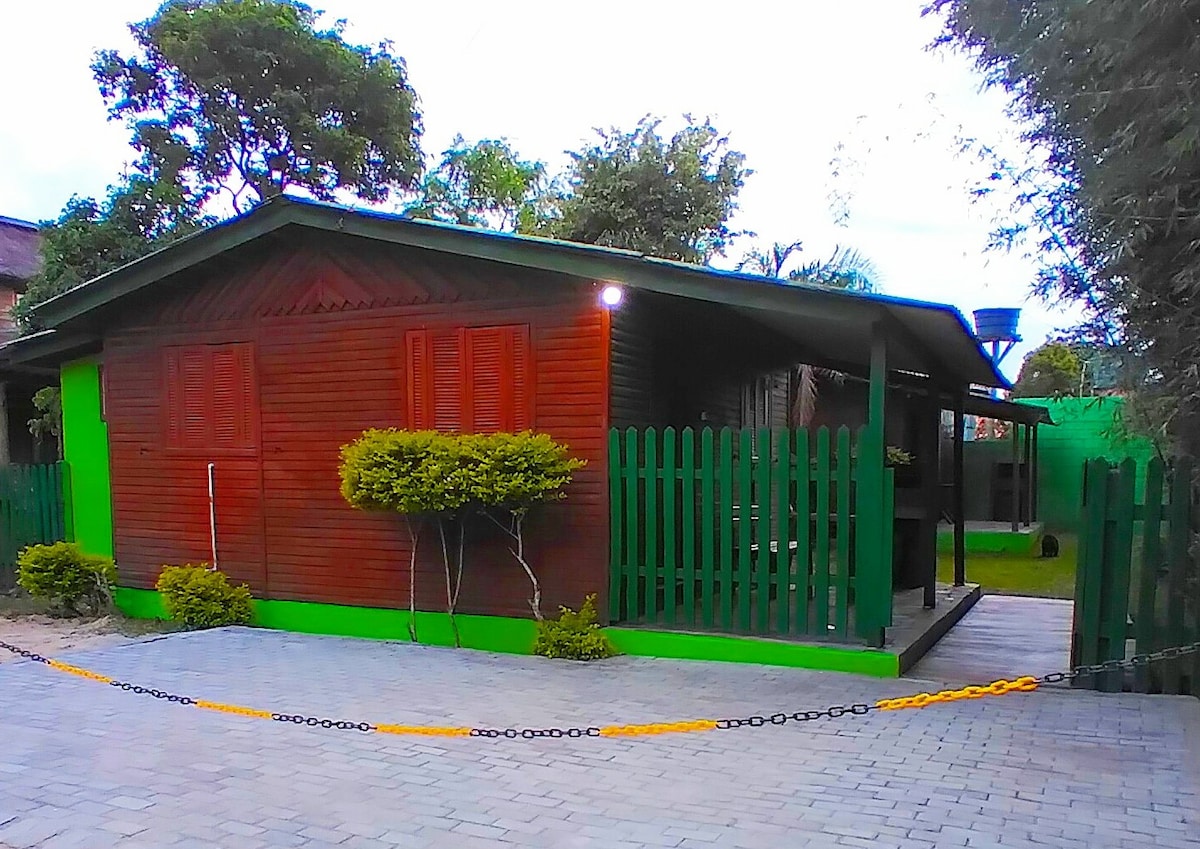 A charming wooden house features a distinctive green trim and shutters. A low fence surrounds the property, complemented by neatly trimmed shrubs. The pathway is paved with rectangular stones, leading to a front porch that invites relaxation in a natural setting.