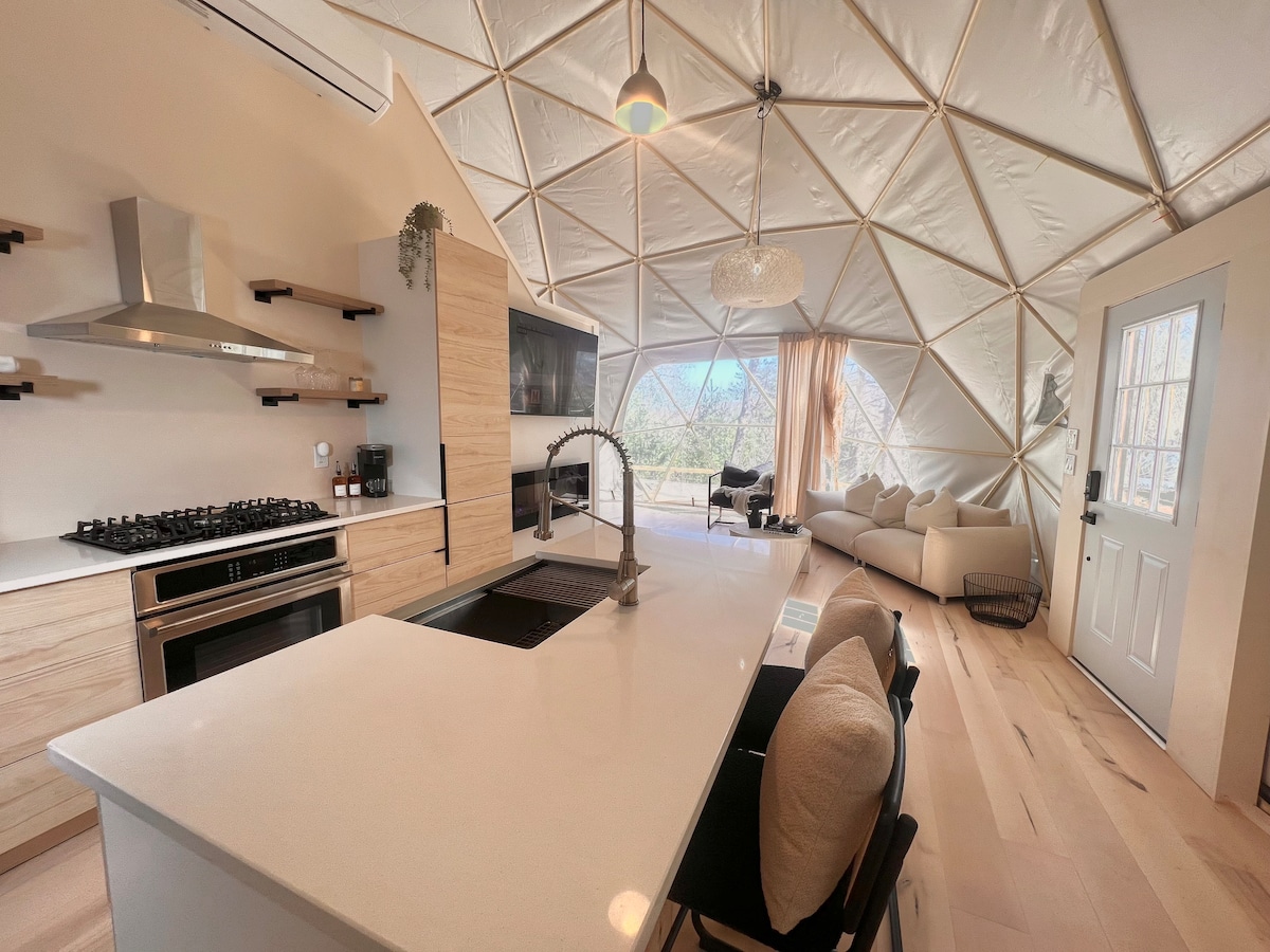 An open-concept kitchen features sleek cabinetry and modern appliances, including a stainless steel stove and a large island with a contemporary sink. A cozy seating area is visible through the large windows, allowing natural light to fill the space and providing views of the outdoors.