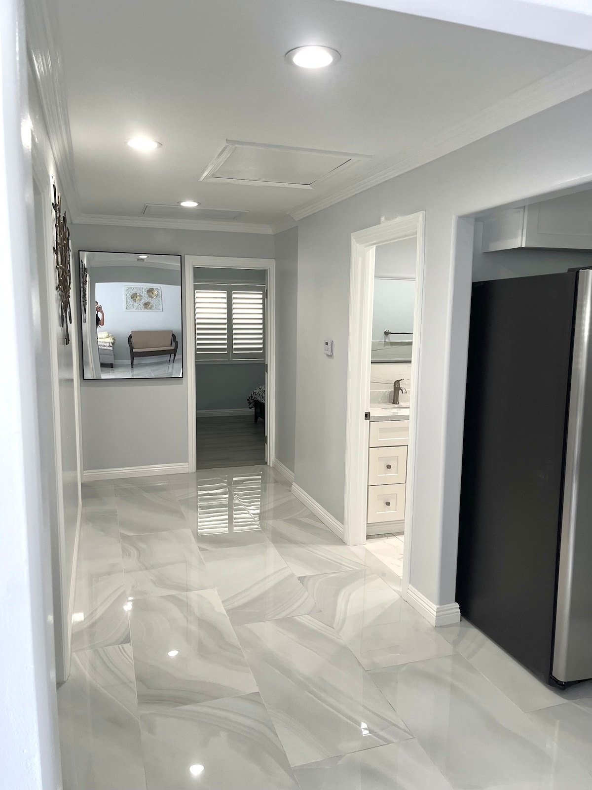 A well-lit hallway features sleek marble flooring and crisp white walls. Doors lead to various rooms, and a glimpse of an adjacent area reveals a dining table in the background. Natural light enhances the bright atmosphere, creating an inviting transition space.