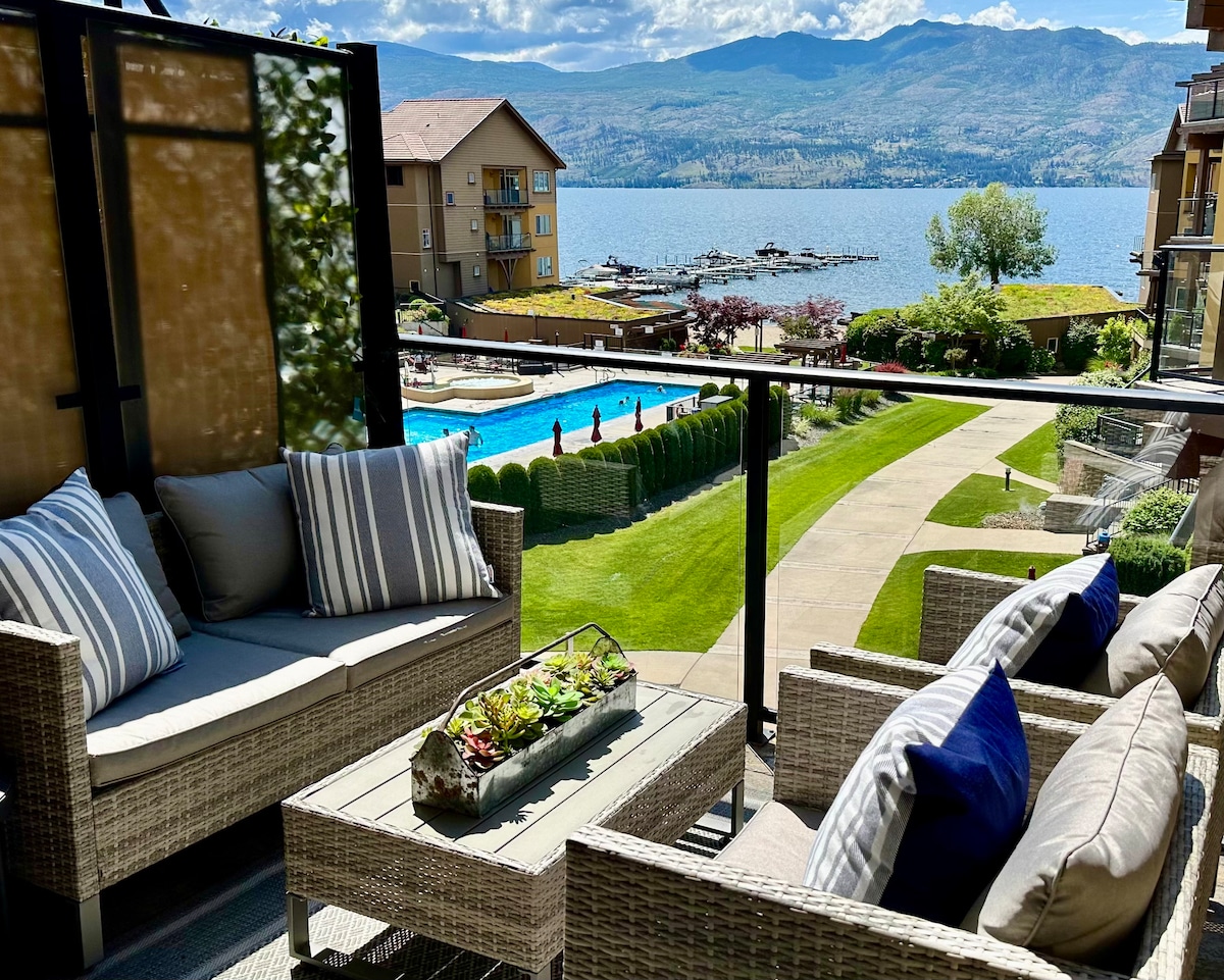 An upper-level patio is furnished with a conversation set featuring plush, striped cushions. A view of the resort's pool and the lake is visible beyond lush greenery, framed by surrounding accommodations and mountains in the distance.