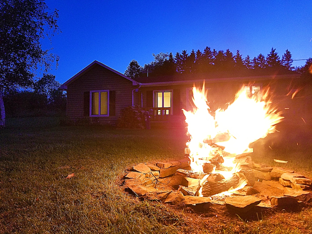 A vibrant fire is surrounded by a circular stone perimeter, casting a warm glow. In the background, a ranch house is visible with large windows reflecting the evening light, framed by a tree line under a deep blue sky.
