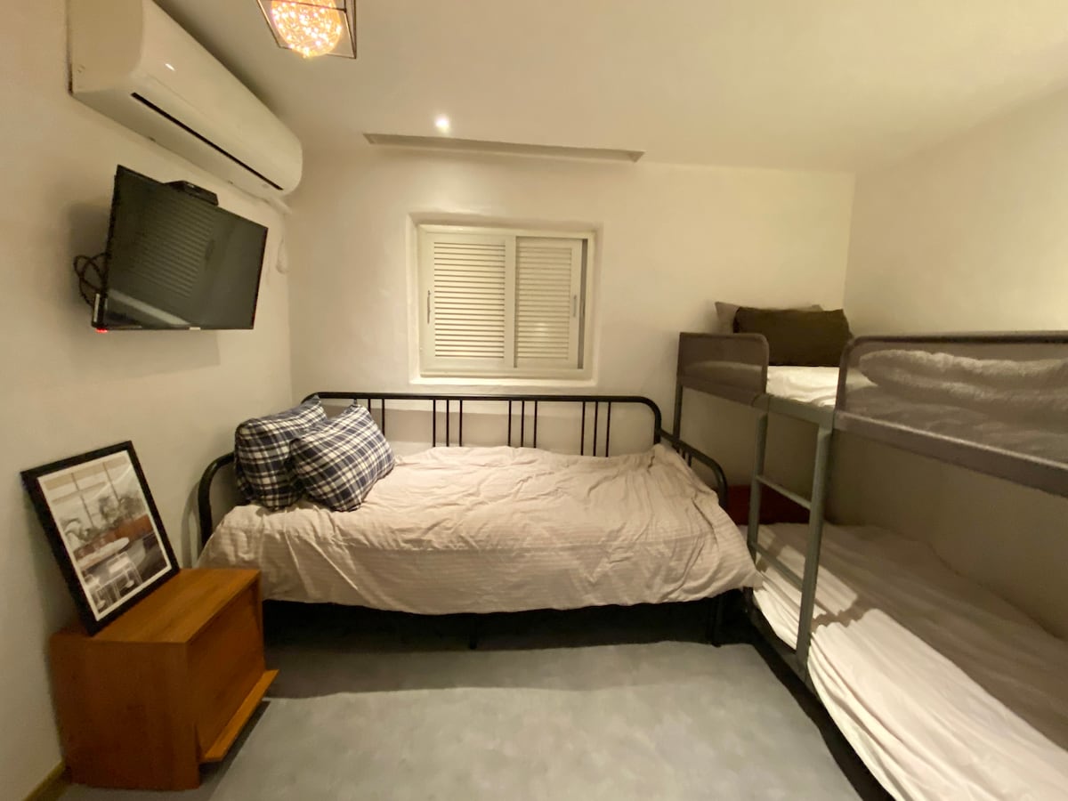 A shared room features a twin over full bunk bed, alongside a sofa bed which expands into a queen size. A wall-mounted television and a small bedside table add functionality to the space. Natural light enters through a window with closed blinds.
