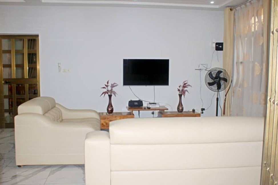 A modern living area is presented, featuring light-colored, spacious seating. A sleek television is mounted on the wall above a wooden table. A standing fan is also visible, providing comfort, with soft natural light entering through partially closed curtains.