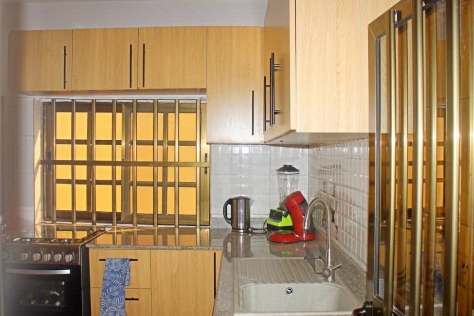 The modern kitchen features wooden cabinetry and a countertop with a stainless steel sink. A blender and kettle are positioned next to the sink, while a gas stove is installed against the wall. Protective bars on the window allow natural light into the space.