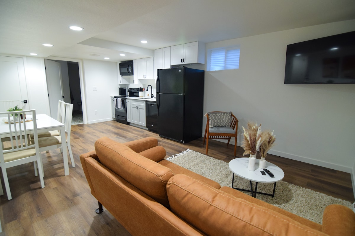 A stylish living area is presented, featuring a comfortable brown sofa and a small coffee table. A dining table with four chairs is positioned nearby. The kitchen area includes modern appliances and cabinetry. Natural light is provided by recessed lighting and a window.