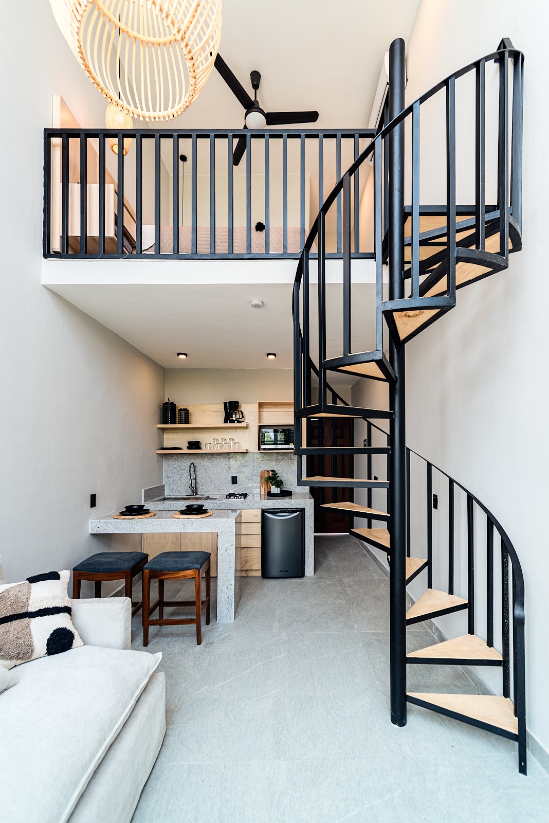 A modern loft interior features a spiral staircase leading to an upper level. A light-colored couch and two wooden stools complement the kitchen area, which includes minimalistic cabinetry and countertop appliances. Soft lighting highlights the open space, enhancing a sense of comfort and functionality.