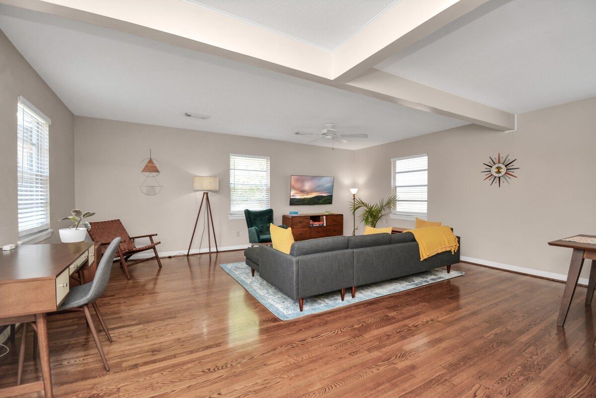 A spacious living area is presented, featuring a large sectional sofa with a yellow throw blanket. A stylish coffee table sits on a decorative rug, complemented by a mid-century desk and lamps. Natural light filters through multiple windows, enhancing the wood flooring.