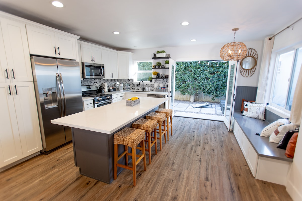A modern kitchen features white cabinetry and stainless steel appliances. A large island with seating provides space for meals, while a window offers a view of the outdoor area. Bright lighting and wooden flooring contribute to a warm ambiance.