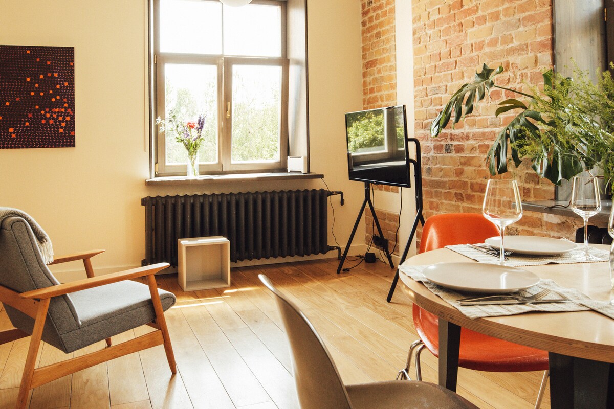 A bright living space features hardwood flooring, exposed brick walls, and large windows allowing natural light. A dining table for four sits beside stylish chairs, with a comfortable armchair positioned nearby. A TV on a stand and floral décor enhance the cozy atmosphere.