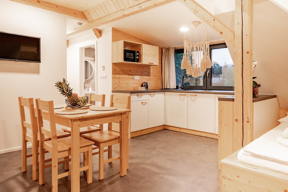 A functional kitchen area features wooden cabinetry and a microwave, with a dining table set for four. Natural light fills the space through the window, and a cozy seating area is visible in the background, creating a welcoming atmosphere.