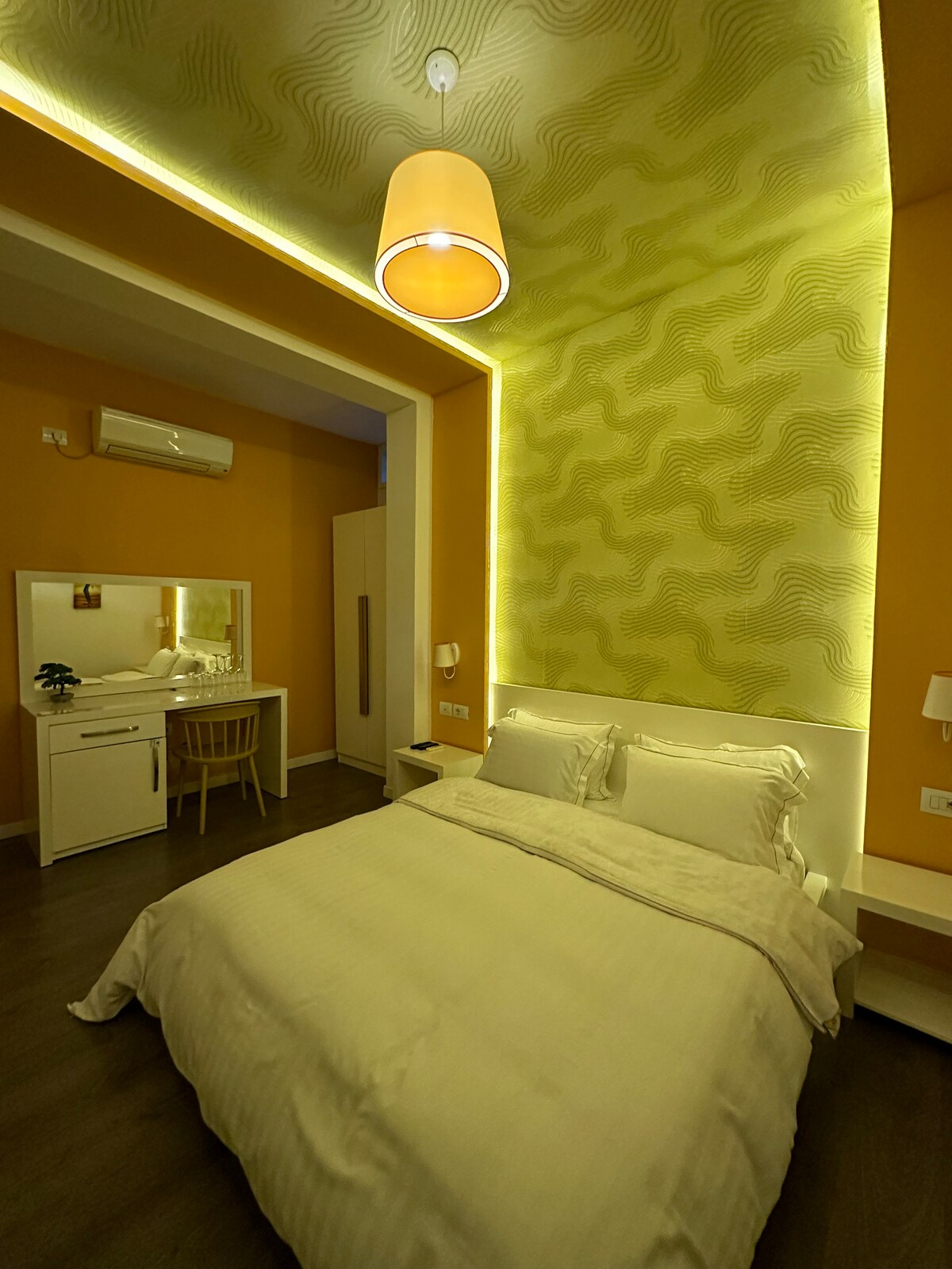 A cozy sleeping area is highlighted by warm yellow and green accents. Soft bedding is arranged neatly on the spacious bed, while a small vanity area with a chair is positioned nearby. An overhead light provides a gentle illumination, enhancing the room's inviting ambiance.