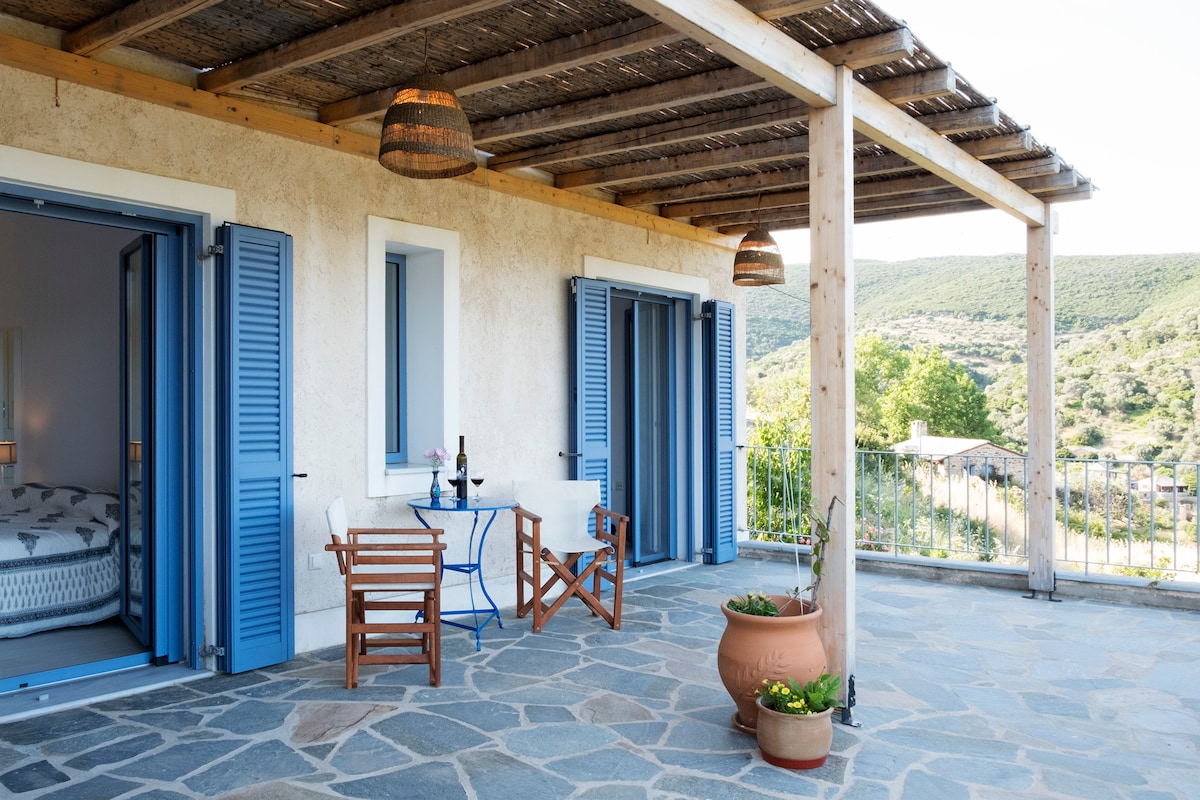 A spacious terrace is depicted, featuring wooden chairs and a blue table. Light blue shutters frame the entrance to the interior room. The roof is supported by wooden beams, with woven baskets hanging overhead. The scenic hills of Zarakes Village are visible in the background.