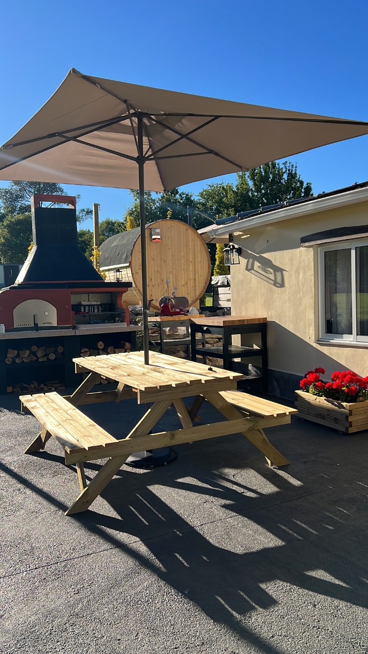 A wooden picnic table is positioned beneath a large umbrella, surrounded by vibrant red flowers in planter boxes. A barbecue grill and a large circular wood storage unit are visible in the background, with clear blue skies overhead.
