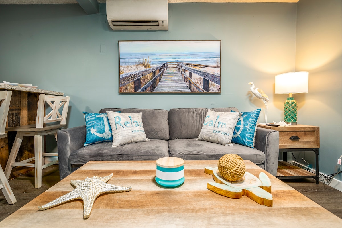 A comfortable living area features a gray sofa adorned with decorative pillows. A wooden coffee table displays a starfish and a carved wooden piece. A serene beach scene is visible on the wall, complemented by a nightstand with a lamp and a green decorative object.