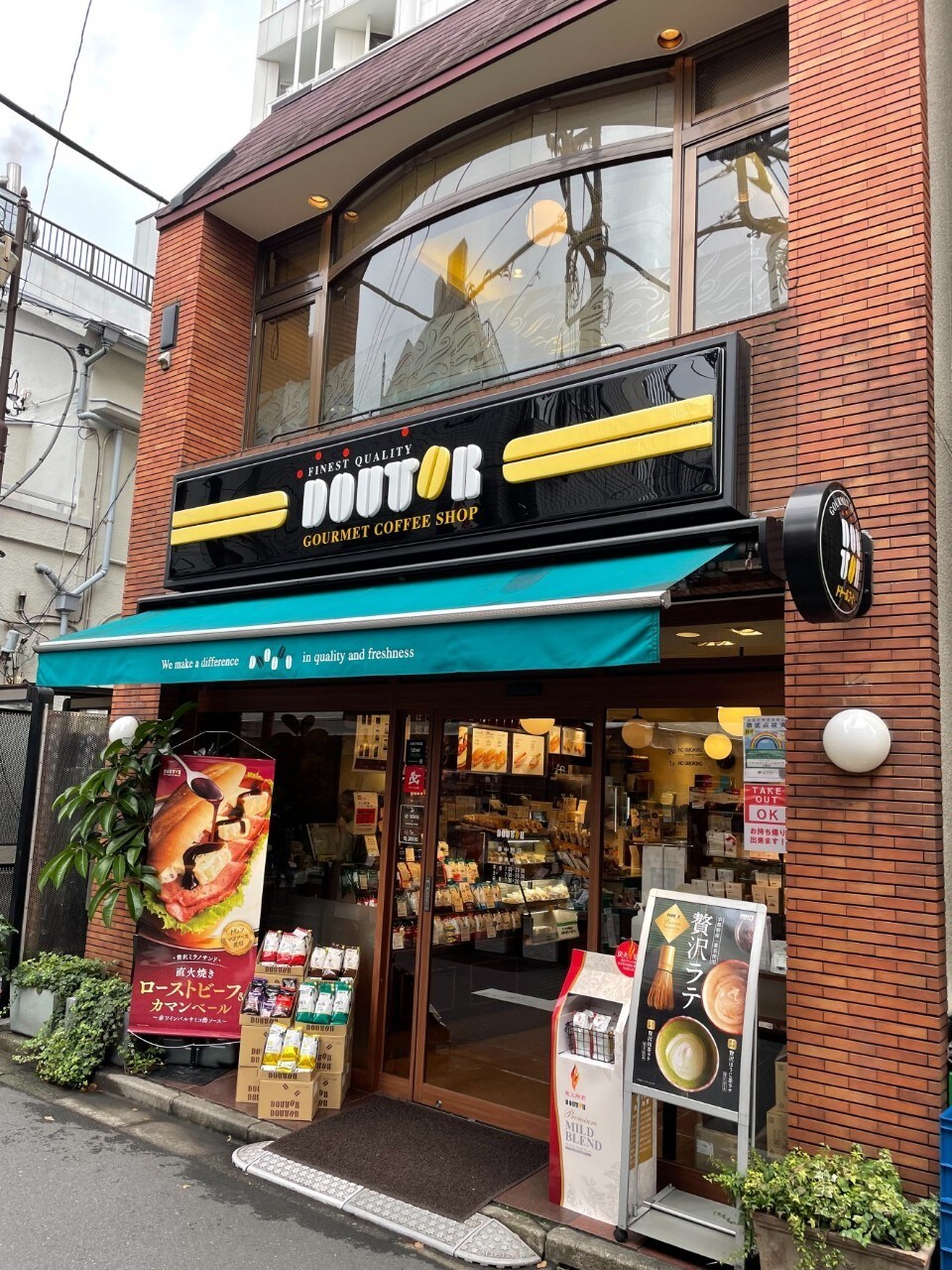 A coffee shop entrance is visible, featuring a dark sign displaying the shop's name in bold letters. An awning in teal covers the entrance, and a display of various food items is arranged outside. The brick exterior adds to the urban setting.