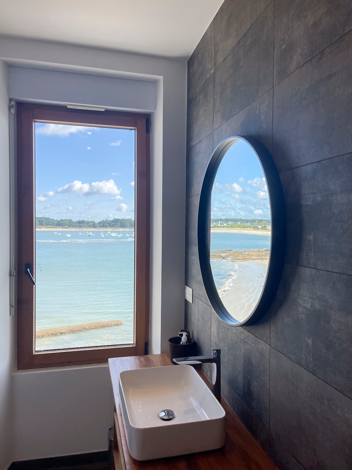 A modern bathroom features a circular mirror above a minimalist sink. A large window allows natural light to fill the space, providing a view of the tranquil sea and distant shoreline, enhancing the sense of openness and connection to the outdoor environment.