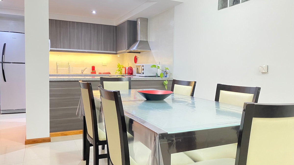 The dining area is furnished with a dark wooden table surrounded by light-colored chairs. A sleek kitchen is visible in the background, featuring modern appliances and a subtle backsplash. Soft lighting highlights the countertops, creating a welcoming environment for meals and gatherings.