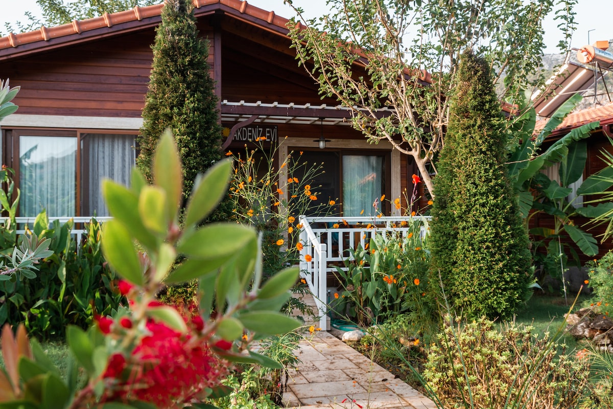 A wooden cabin is framed by lush greenery and vibrant flowers, with a stone pathway leading to the entrance. Large windows allow for natural light, while the surrounding plants enhance the tranquil outdoor setting.