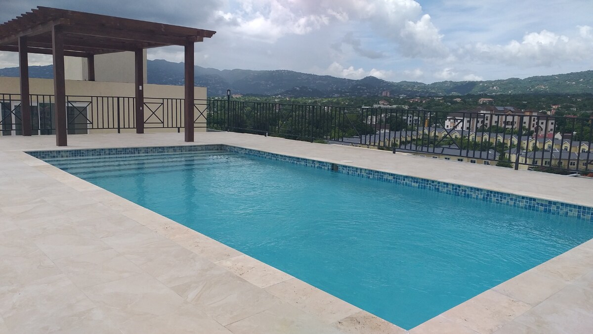 A rooftop pool features clear blue water, surrounded by a spacious tiled deck and bordered by a black railing. A shaded pergola is present at one end, offering a relaxed space to unwind while enjoying panoramic views of the city and mountains.