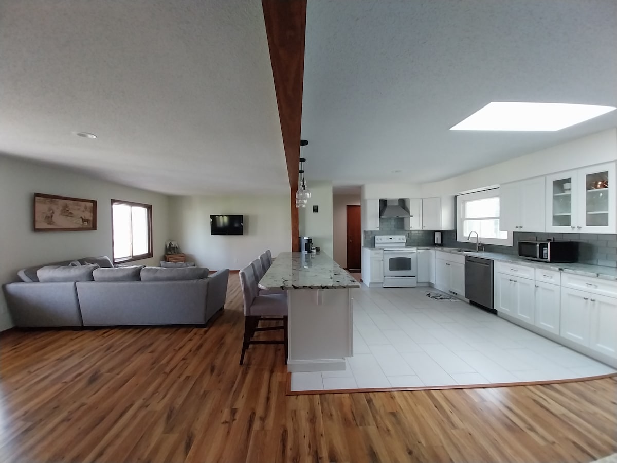 An open-concept living space showcases a modern kitchen with white cabinetry and stainless steel appliances. A large island with seating accommodates multiple guests. The cozy living area features a comfortable sectional sofa and a wall-mounted television, complemented by natural light from multiple windows.