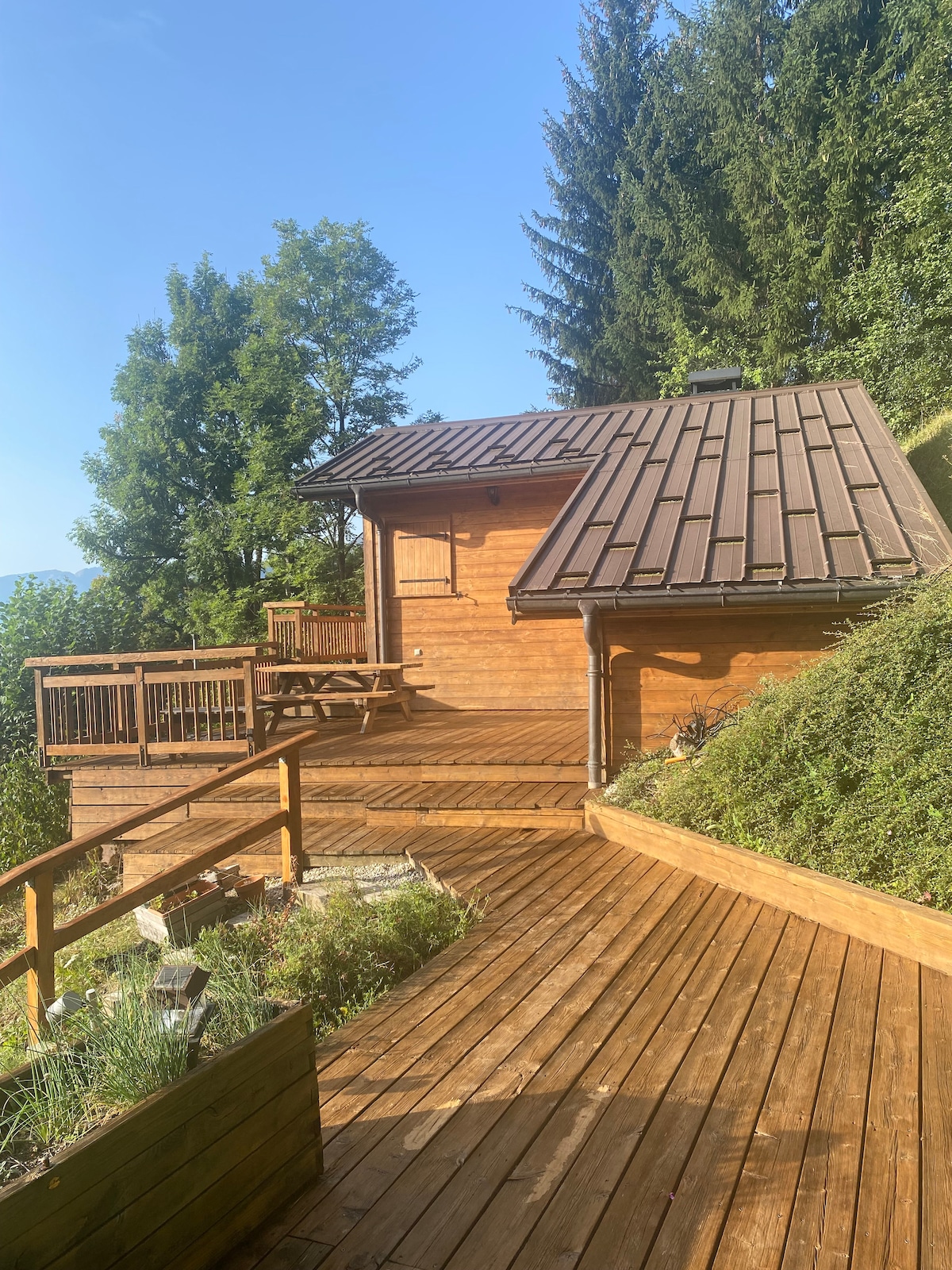 A wooden chalet is set against a backdrop of greenery, featuring a sloped roof and a spacious wooden deck. The deck includes a picnic table and is surrounded by a path made of wooden planks. Lush trees provide a natural border around the property.