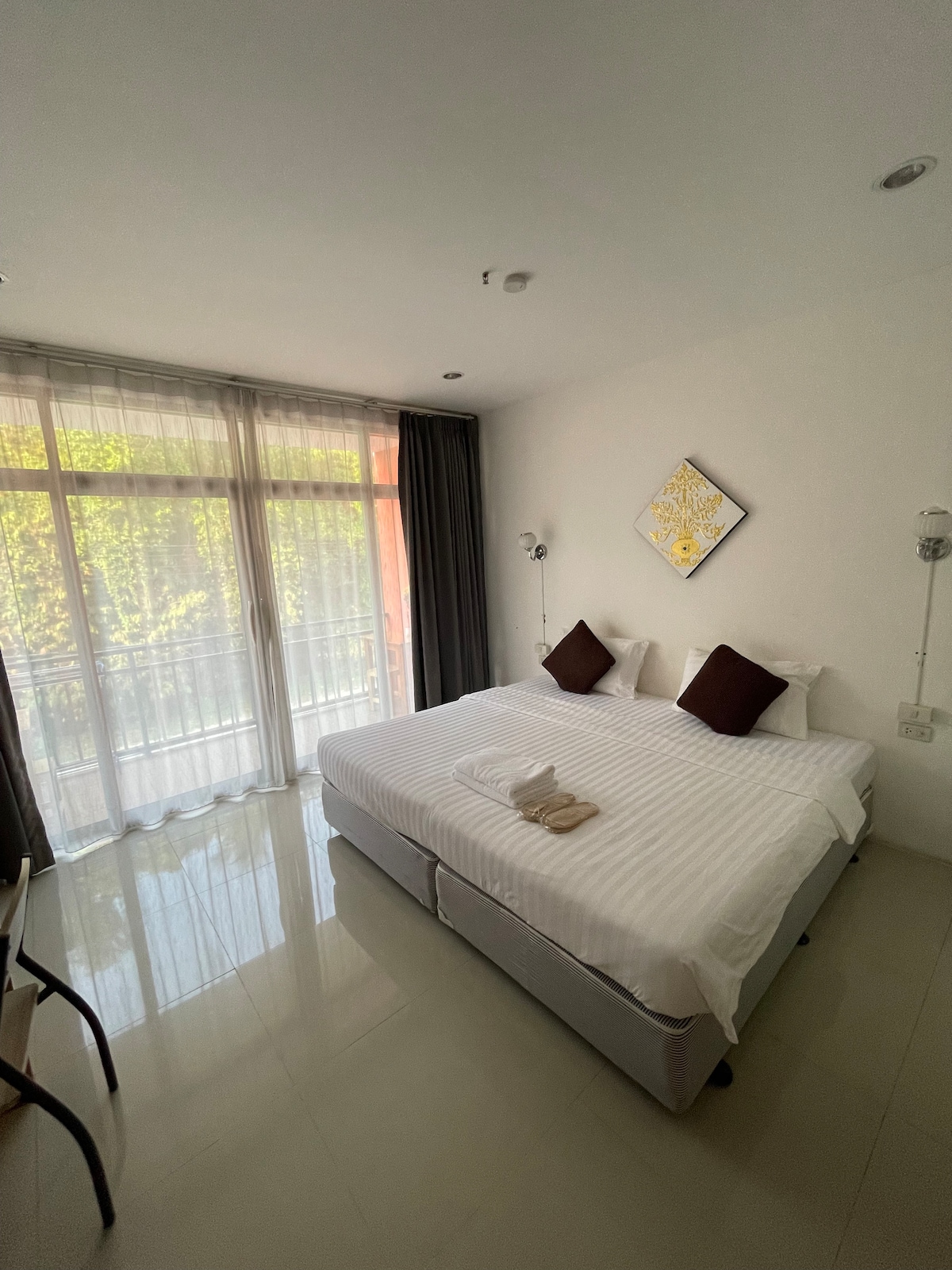 A bright and airy room features a spacious bed with crisp white linens and dark accent pillows. Natural light filters through large windows, which are framed by sheer curtains. Minimalist decor includes a simple artwork piece above the bed and a small desk nearby.