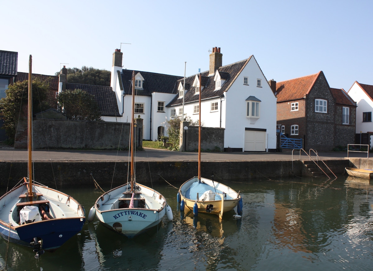 Three small boats are anchored closely together in the calm water, showcasing white and blue hulls. The shore features several charming coastal buildings with varied roof styles, including peaked roofs and chimney stacks, while the clear sky adds to the serene atmosphere.