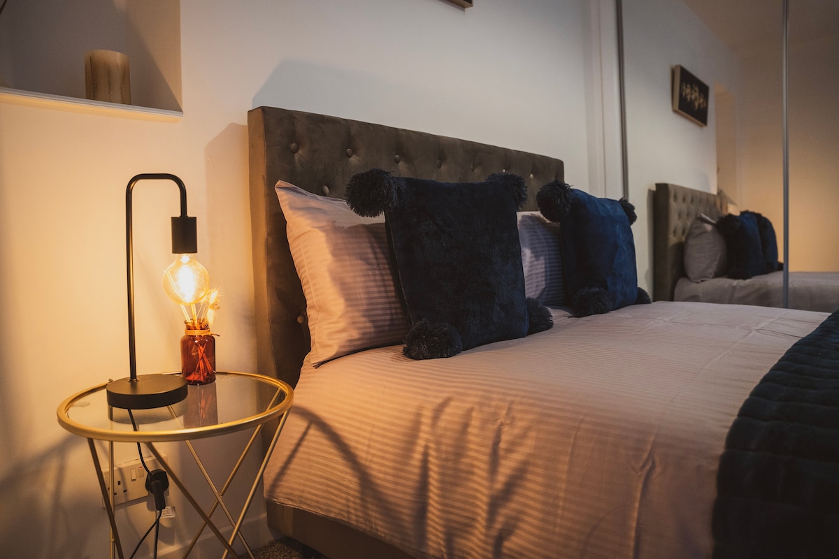 A bedroom is shown with a double bed featuring plush pillows and a soft blanket. A stylish bedside lamp sits on a small round table beside the bed, casting a warm glow. The fitted wardrobe is visible in the background, enhancing the room's spaciousness.