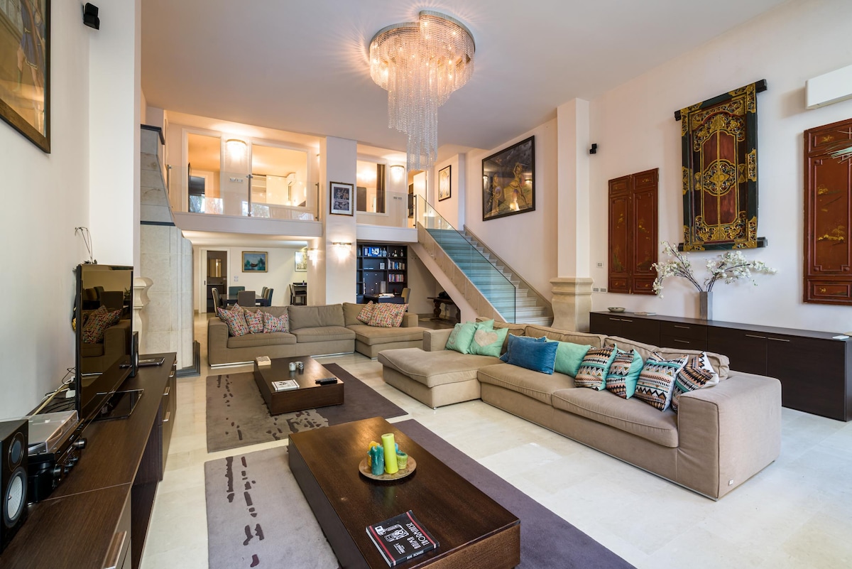 A spacious living area is displayed, featuring multiple seating arrangements with plush sofas and decorative cushions. A large chandelier hangs from the ceiling, and a sleek entertainment unit is positioned against the wall. The room is illuminated by soft lighting, creating an inviting environment.