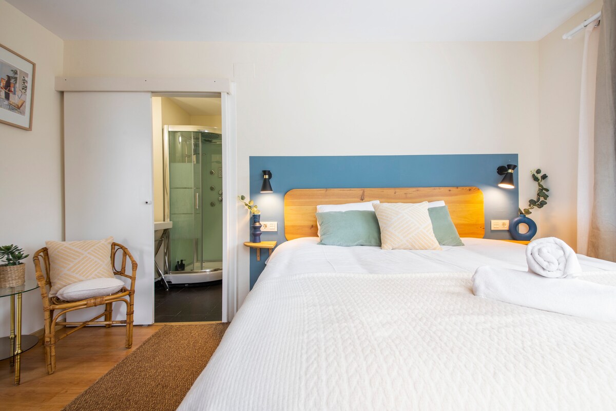 A cozy bedroom features a spacious bed with neutral-toned bedding and decorative pillows. A rattan chair sits beside the bed. Natural light enters through a nearby window, while an ensuite bathroom is visible through an open door.
