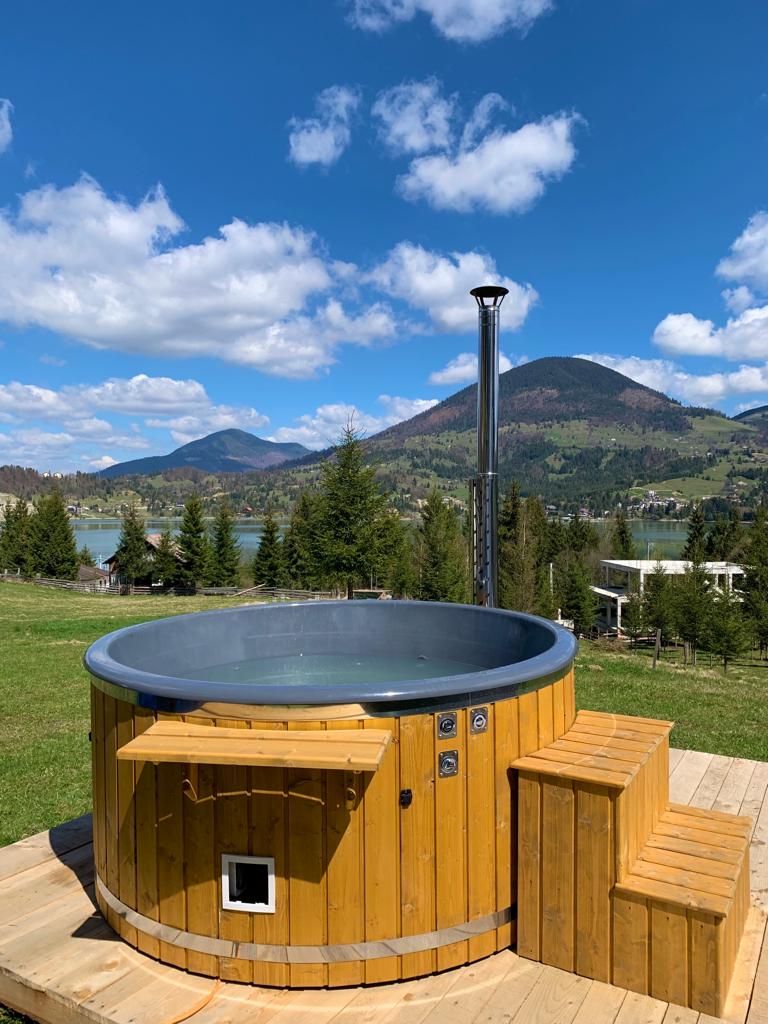 A wooden hot tub is positioned on an outdoor deck, surrounded by a lush green landscape. Rolling hills and mountains create a scenic backdrop, while clear blue skies with scattered clouds enhance the peaceful atmosphere.