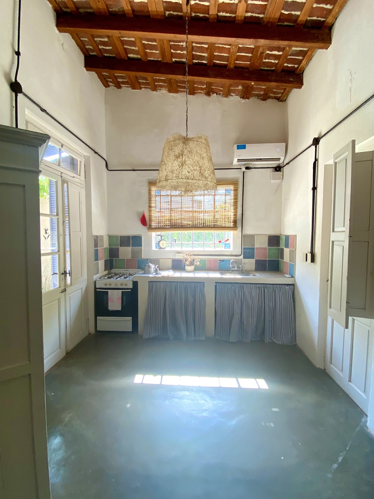 A bright kitchen area features high wooden ceilings and a large hanging light fixture. The space includes a small stove and sink, with cabinets displaying patterned tile accents. Natural light floods in from a window, creating an open and airy atmosphere.