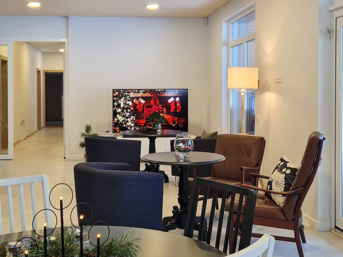 A well-furnished living area features a television with festive decorations. A round table is surrounded by three chairs and a reclining armchair, creating a cozy setting. Natural light enters through large windows, contributing to a bright and welcoming ambiance.