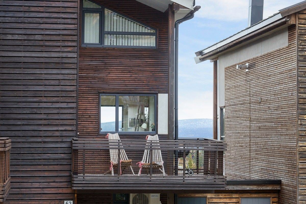 A balcony area showcases two wooden deck chairs with soft throw blankets, providing a relaxing spot to enjoy outdoor views. The building's modern wooden facade contrasts with the neighboring structure, enhancing the contemporary architectural design.
