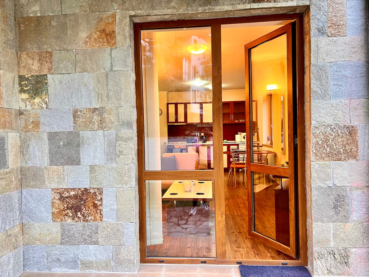 An entrance view features a wooden framed door open to reveal a stylish interior. A spacious living area is visible, including a dining table and a well-equipped kitchen. Natural stone exterior provides a textured backdrop, while warm lighting creates an inviting ambiance.