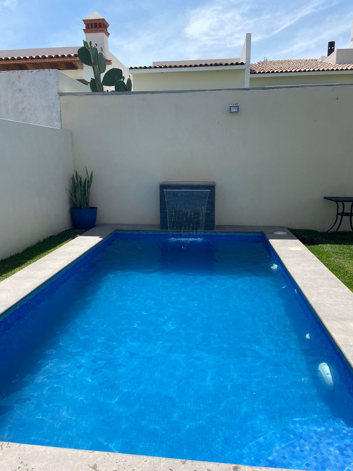 A private rectangular pool is featured in a tranquil outdoor space, with clear blue water and a small waterfall. Surrounding the pool, a well-maintained lawn and a simple table set against the wall add to the peaceful ambiance.