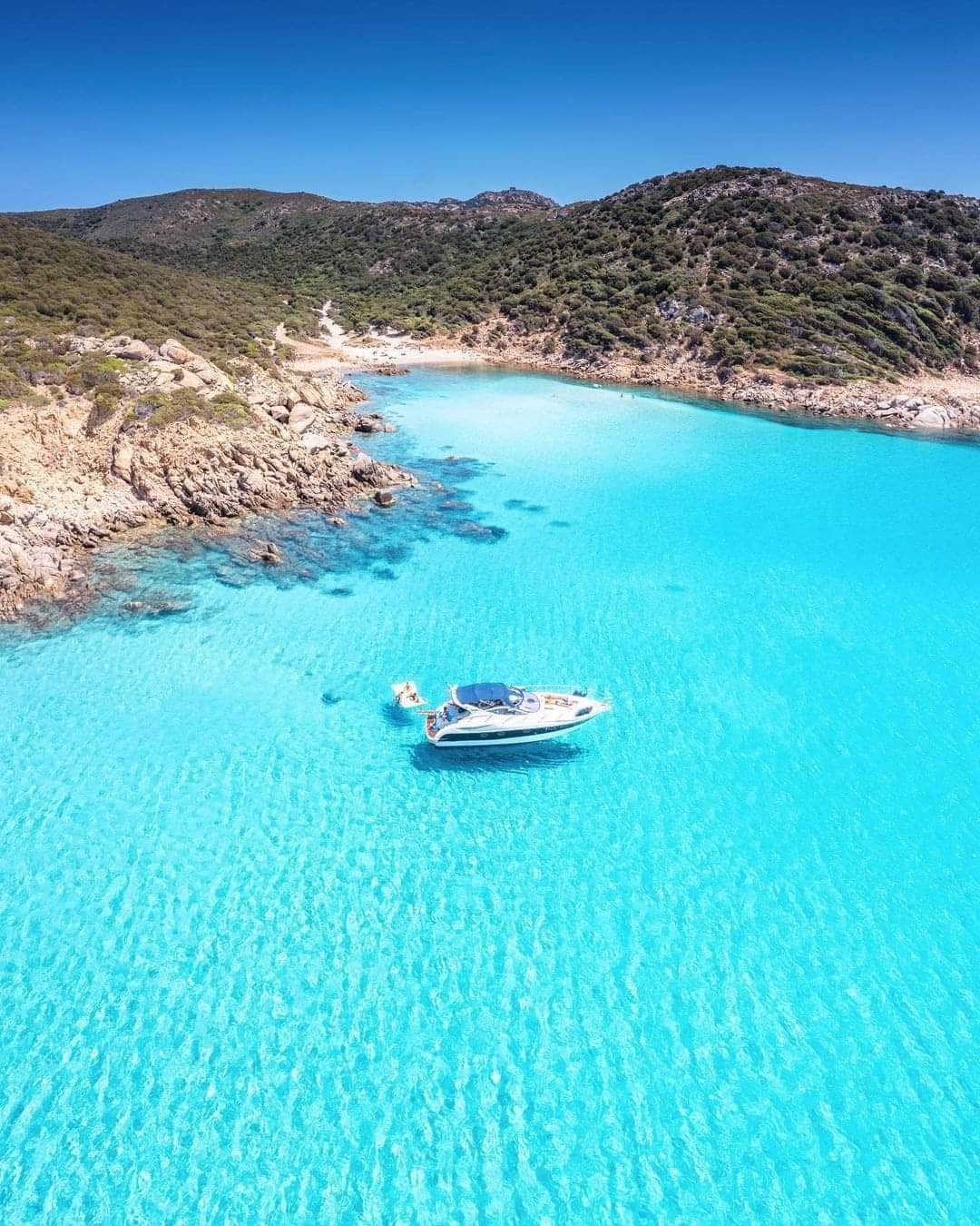 A clear turquoise sea is depicted, with gentle waves lapping against the rocky shoreline. A boat is anchored in the water, surrounded by lush green hills that rise in the background under a bright blue sky.