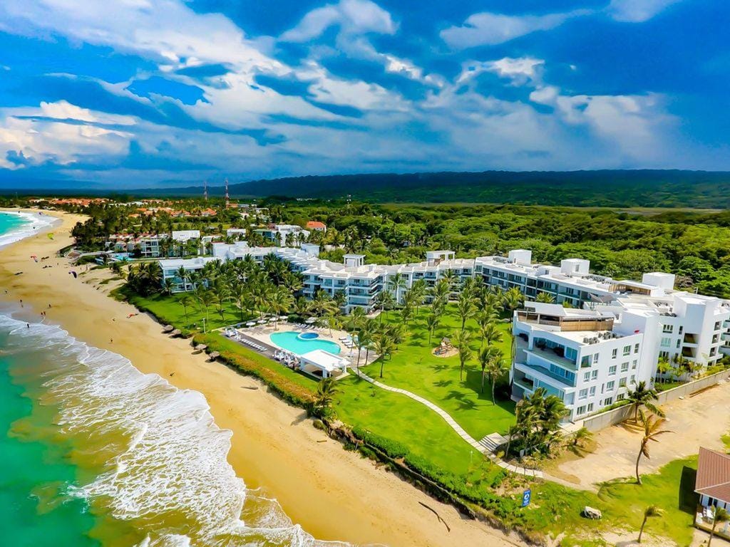 An aerial view captures the luxury beachfront condo complex nestled along the coastline. The property is surrounded by lush greenery and features several swimming pools. The sandy beach gently curves along the water's edge, inviting moments of relaxation by the ocean.
