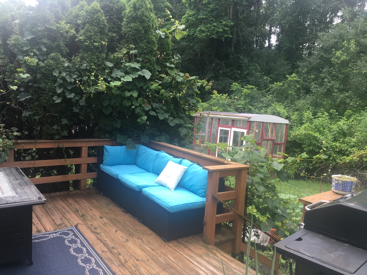 A wooden deck is adorned with a sectional seating arrangement featuring vibrant blue cushions. Lush greenery surrounds the area, providing a sense of privacy. A grill is positioned nearby, complemented by a small table adorned with a plant. A colorful shed is also visible in the background.