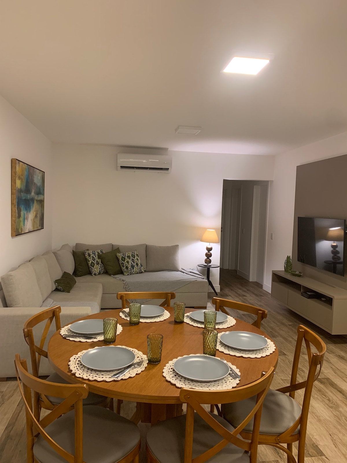 A cozy living area features a light grey sofa with decorative cushions, complemented by a round wooden dining table set for six. Green glassware adds a touch of color, while a modern TV and ambient lighting enhance the inviting space.