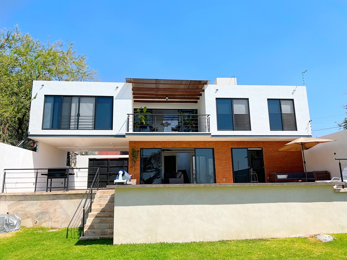 The modern two-story house features large windows and a spacious terrace. A well-manicured green lawn surrounds the property, while the upper balcony offers shaded seating beneath a roofed area. Outdoor furniture is positioned on the terrace, highlighting the home’s inviting layout.