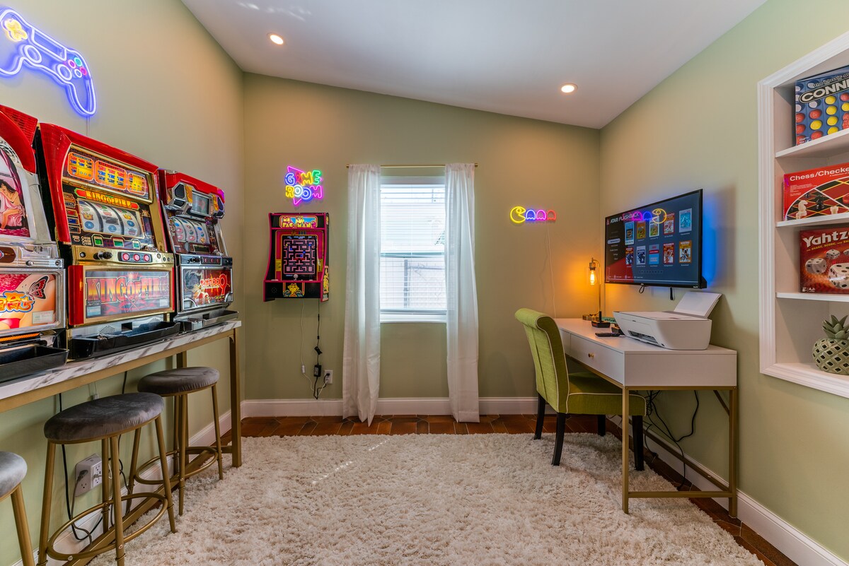 A game room is highlighted by colorful neon signs and classic arcade machines. Two slot machines and an Atari game are positioned against the walls. A small desk with a wireless printer is situated near a window, providing natural light. A soft, white area rug covers the floor.