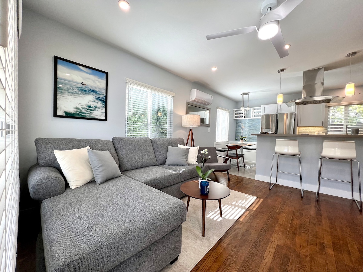 A spacious living area features a grey L-shaped sofa topped with decorative pillows. A round coffee table rests in front, accompanied by a floor lamp and artwork on the wall. Natural light filters through multiple windows, illuminating the open kitchen in the background.
