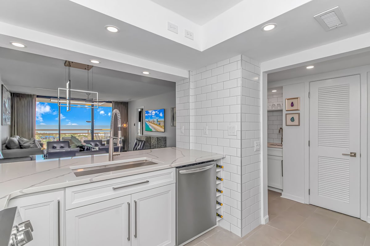 A modern kitchen is featured with clean white cabinetry and a spacious countertop, equipped with a sleek sink and contemporary faucet. The background reveals an open area leading to a living space with large windows showcasing an ocean view. Additional rooms are indicated by a doorway to the right.